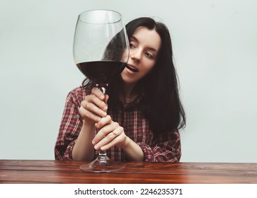 A young girl in a red plaid shirt gently holds a large glass of red wine and looks at him in surprise. The concept of alcohol abuse, alcoholism, hangover, loneliness and depression. Grey background. - Shutterstock ID 2246235371