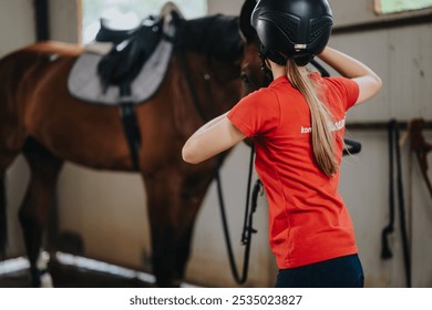 Young girl in a red shirt preparing a horse for riding in a stable. She is wearing a helmet and adjusting it. - Powered by Shutterstock