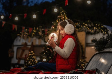 Young girl in red vest and earmuffs admires glowing snow globe, sitting near Christmas tree with warm holiday lights in the background. Back yard of Camping van - Powered by Shutterstock