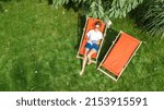 Young girl relaxes in summer garden in sunbed deckchair on grass, woman reading book outdoors in green park on weekend, aerial drone view from above