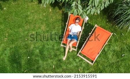 Young girl relaxes in summer garden in sunbed deckchair on grass, woman reading book outdoors in green park on weekend, aerial drone view from above