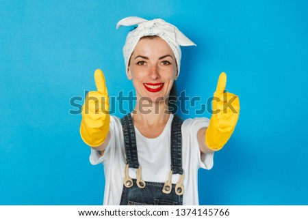 Young girl with a scarf on her head and yellow rubber gloves showing a gesture of thumbs up, super, like on a blue background. Concept of cleaning and cleaning service, high quality.