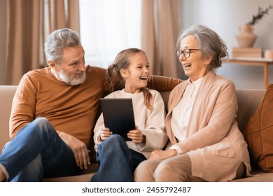 A young girl, seated between her grandparents, shares a joyful moment with them on a comfortable couch, using digital tablet together at home - Powered by Shutterstock