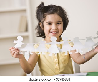 A Young Girl Is Seated At A Desk And Is Holding Up Paper Dolls.  She Is Smiling At The Camera.  Horizontally Framed Shot.