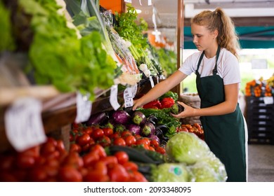Young girl seller in uniform working in supermarket at her first job, checking organic eggplants - Shutterstock ID 2093348095