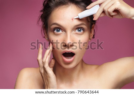 Young girl in shock of her acne. Photo of ugly girl with problem skin applying treatment cream on pink background. Skin care concept