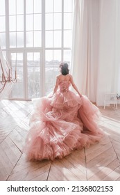 young girl with short black hair in pink princess dress with train is standing from the back and looking cute near huge panoramic white window background at sunny day. fashion concept, free space - Shutterstock ID 2136802153