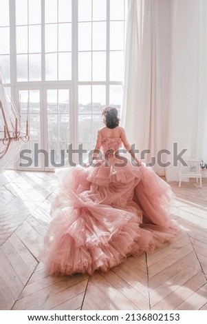 young girl with short black hair in pink princess dress with train is standing from the back and looking cute near huge panoramic white window background at sunny day. fashion concept, free space
