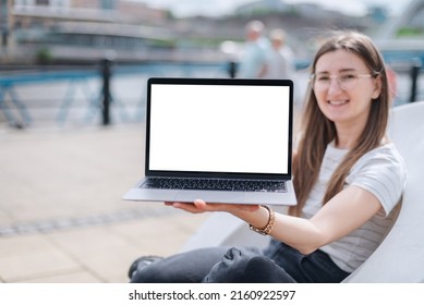 A Young Girl Shows Off A Laptop With A Blank Screen While Sitting In A Modern Plastic Chair On The Waterfront. A Laptop Mockup With A Blank White Screen.