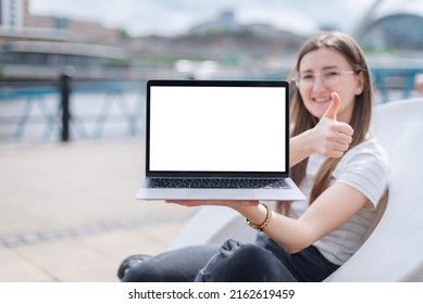 A Young Girl Shows Off A Laptop With A Blank Screen While Sitting In A Modern Plastic Chair On The Waterfront. A Laptop Mockup With A Blank White Screen.