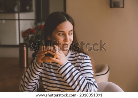 A young girl sits in a chair, looks away and drinks a hot drink. A woman holds a cup in her hands and rests after a working day and looks out the window. Cup of tea or cofee. 