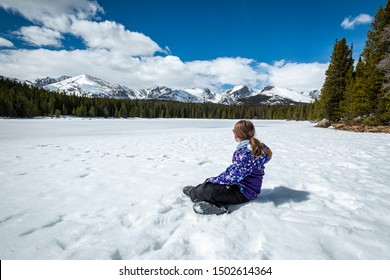 Young Girl Sits On Frozen Lake On A Beautiful Sunny Winter Afternoon Looking At Snow Covered Mountains In The Distance.  Bierstadt Lake In Rocky Mountain National Park, Colorado