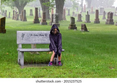 Young girl sits on her a family bench in a grave yard with a hoodie not keeping her dry in the rain, looking down contemplating looking sad.