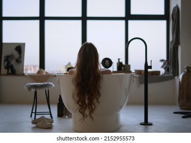A young girl is sitting in the bathtub, rear view, frame from the back. Large bathroom with windows to the floor. Girl with long hair.
 - Powered by Shutterstock