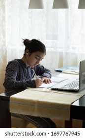 Young Girl Sitting At The Dining Table With Laptop At Home Schooling, Online Virtual Classroom Video Conference, Distant Education. Active Participation At The Lesson With Earphones, Doing Homework.