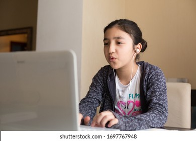 Young Girl Sitting At The Dining Table With Laptop At Home Schooling, Online Virtual Classroom Video Conference, Distant Education. Active Participation At The Lesson With Earphones, Doing Homework.