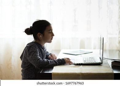 Young Girl Sitting At The Dining Table With Laptop At Home Schooling, Online Virtual Classroom Video Conference, Distant Education. Active Participation At The Lesson With Earphones, Doing Homework.