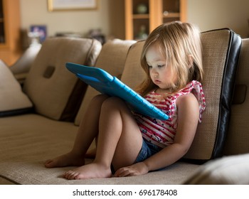Young girl sitting at home on settee and using a child's tablet touch screen computer during home schooling due to closure of schools and pre-schools by coronavirus