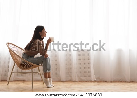 Young Girl Sitting In Modern Chair, Enjoying Coffee In Front Of Window, Side View