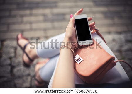 Young girl sitting on a bench with with fashionable leather bag using her empty copy space white mobile phone. Summer vintage technology background.