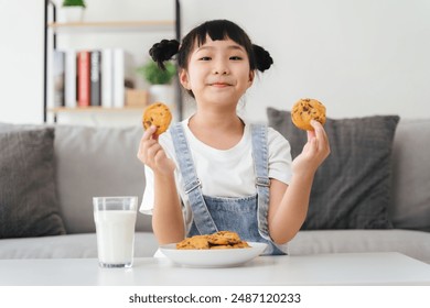 A young girl is sitting on a couch with a glass of milk and a plate of cookies. She is smiling and holding up the cookies - Powered by Shutterstock