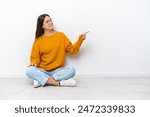 Young girl sitting on the floor isolated on white background pointing finger to the side and presenting a product