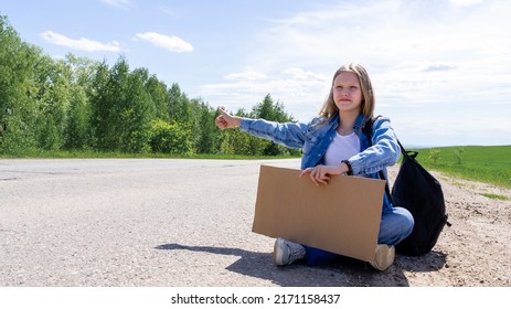A young girl is sitting on the road with a sign in her hands, hitchhiking around the country. The girl is trying to catch a passing car for a trip. A girl with a backpack went on vacation hitchhiking