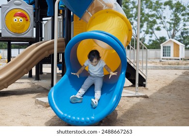 A young girl is sitting on a sky blue slide, smiling and waving. Concept of joy and excitement, as the girl is enjoying her time at the playground - Powered by Shutterstock