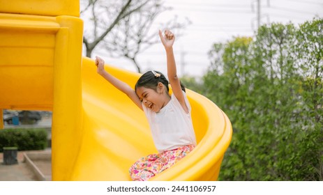 A young girl is sitting on a yellow slide, smiling and waving. Concept of joy and excitement, as the girl is enjoying her time at the playground - Powered by Shutterstock