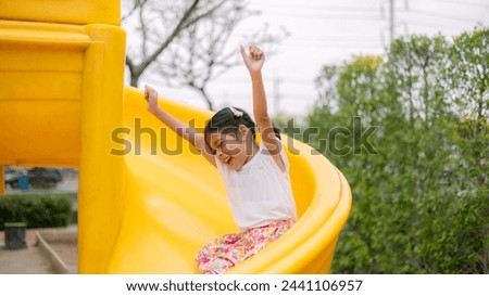 A young girl is sitting on a yellow slide, smiling and waving. Concept of joy and excitement, as the girl is enjoying her time at the playground