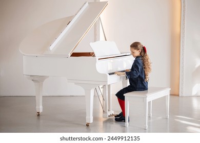 A young girl is sitting at a piano, playing a song. The piano is white and has a black and gold trim. The girl is wearing a blue dress and a red bow. Scene is one of innocence and joy - Powered by Shutterstock