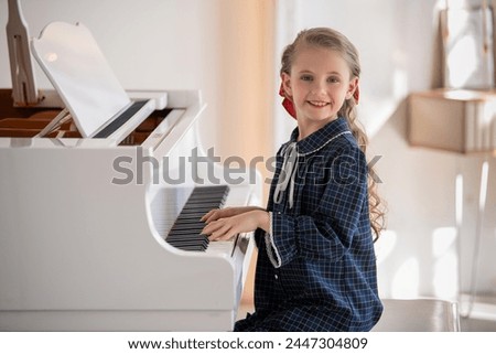 A young girl is sitting at a piano, playing a song. The piano is white and has a black and gold trim. The girl is wearing a blue dress and a red bow. Scene is one of innocence and joy