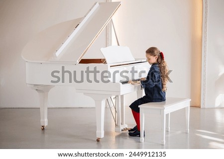 A young girl is sitting at a piano, playing a song. The piano is white and has a black and gold trim. The girl is wearing a blue dress and a red bow. Scene is one of innocence and joy