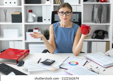 A young girl is sitting at a table in the office, holding a red cup in her hand and working with documents. - Shutterstock ID 1122162674