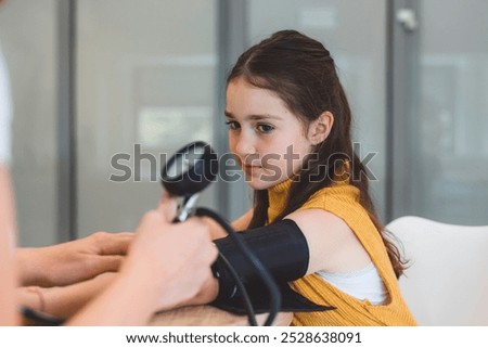 A young girl sitting at a table while a healthcare professional measures her blood pressure. The girl looks calm and attentive, wearing a yellow top. The setting appears to be a medical office or clin