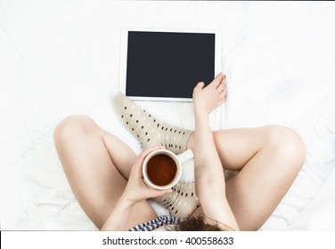 A Young Girl Slim Pretty Bare Legs Sitting On A Bed In The Lotus Position Crossed Legs. Is Using Tablet Holding Cup Of Cocoa. White Sheets. Top View . Spending Time In Bed