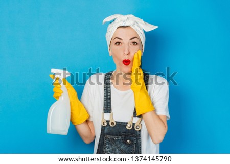 A young girl with a smile and a handkerchief and yellow rubber gloves is holding a spray bottle against a blue background. Cleaning Concept and Cleaning Service.