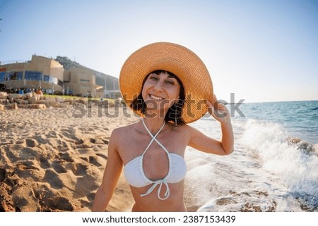 young girl smiling at the camera. Portrait of a beautiful young woman in a straw hat and white bikini on the seashore. Happy girl with black hair enjoying the sun.