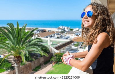 Young girl smiling with sunglasses leaning out on a terrace overlooking the sea. Family summer vacations in beach hotels and resorts. Travel insurance concept. Copy space. - Powered by Shutterstock