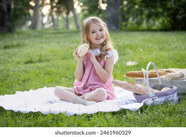 Young girl smiling while holding a bread roll, sitting on a picnic blanket in a lush park. - Powered by Shutterstock