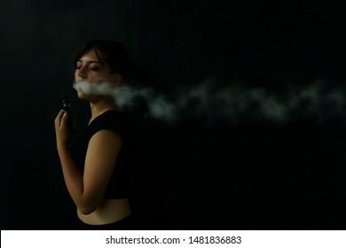 Young Girl Smokes Electronic Cigarette. The Model Vaping A Vaporizer In The Studio.The Face Of Vaping Young Woman At Black Studio. No Smoking Day