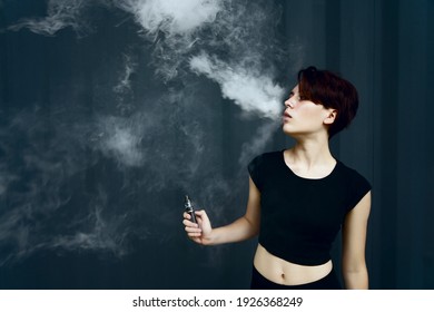 Young Girl Smokes Electronic Cigarette. The Model Vaping A Vaporizer In The Studio.The Face Of Vaping Young Woman At Black Studio. No Smoking Day