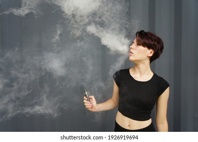 Young Girl Smokes Electronic Cigarette. The Model Vaping A Vaporizer In The Studio.The Face Of Vaping Young Woman At Black Studio. No Smoking Day