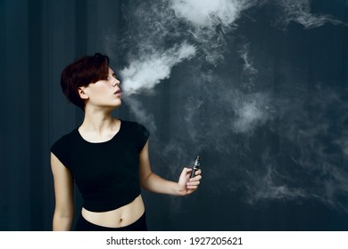 Young Girl Smokes Electronic Cigarette. The Model Vaping A Vaporizer In The Studio.The Face Of Vaping Young Woman At Black Studio. No Smoking Day