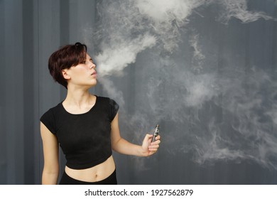 Young Girl Smokes Electronic Cigarette. The Model Vaping A Vaporizer In The Studio.The Face Of Vaping Young Woman At Black Studio. No Smoking Day