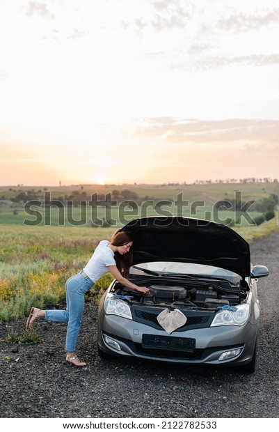 A young girl\
stand near a broken car in the middle of the highway during sunset\
and tries to repair it. Troubleshooting the problem. Car service.\
Car breakdown on the road.