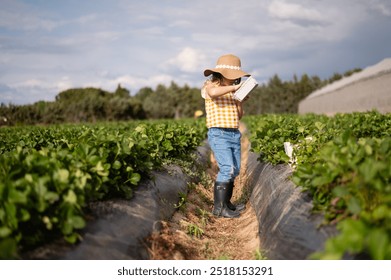 A young girl is standing in a field of strawberries, wearing a straw hat and holding a book - Powered by Shutterstock