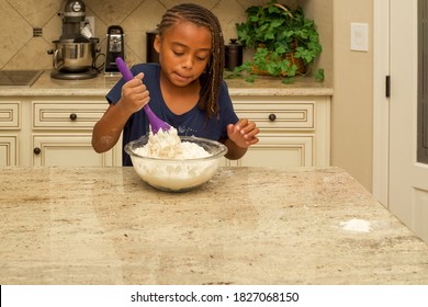 A Young Girl Is Standing At The Kitchen Counter Mixing Dough In A Clear Glass Bowl With A Purple Mixing Spoon.