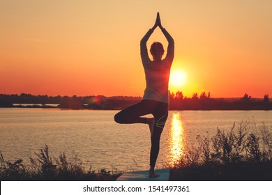 A young girl is standing on the lake at sunset, doing yoga. Stands in a pose of tree a Sathi Yoga. Balance, harmony, balance, concentration, relaxation
