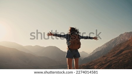 Young girl standing on top of mountain and victoriously raising hands up, looking far away - zennism, freedom, adventure concept 